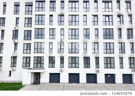 A modern white apartment building features large black-framed windows, several air conditioning units, and garage doors 124655079