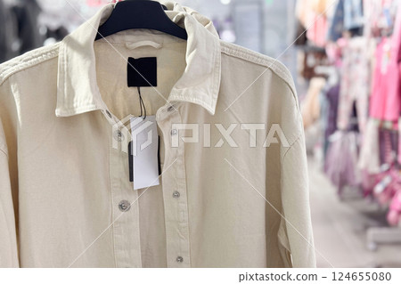 A beige button-up shirt with a blank price tag hangs on a rack in a brightly lit clothing store 124655080