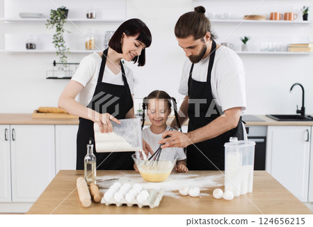 Caucasian family includes father, mother, young daughter, baking in modern kitchen while smiling. Father mixing batter. Mother pouring ingredients. Daughter watching activity. 124656215