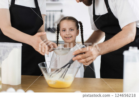 Caucasian family with young girl cooking in bright kitchen. Focus on teaching and teamwork. Includes flour, bowl, whisk engaging girl with smiling face and braided hair. 124656218