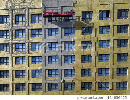 Apartment building facade under renovation with insulation and scaffold. Wall partially covered with gray facade material and yellow insulation panels. Construction site setting 124656455
