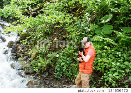 Man wearing cap and sunglasses stands by forest stream, holding black cat Outdoor adventure in lush green woodland, tranquility and connection with nature, Traveling with pet, walking copy space 124656470
