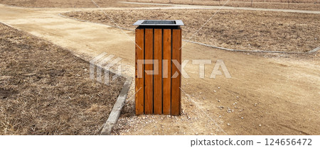 Wooden waste bin stands on a dirt path in an open park setting. Surrounding landscape is dry and grassy, typical of a dry weather season, emphasizing environmental awareness , copy space Wooden waste bin stands on a dirt path in an open park setting. Surrounding landscape is dry and grassy, typical of a dry weather season, emphasizing environmental awareness , copy space 124656472