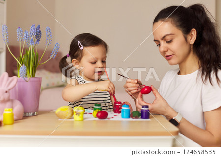 Happy mother and cute little child girl are preparing to celebrate Easter at home. Mom and daughter painting eggs before Easter together. Happy mother and cute little child girl are preparing to celebrate Easter at home. Mom and daughter painting eggs before Easter together. 124658535