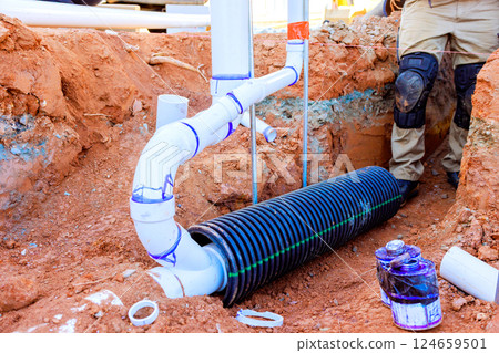 Worker are busy placing black drainage pipe among newly laid white PVC in trench at construction site 124659501