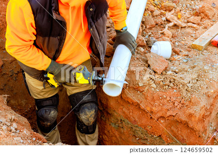 Worker in an orange sweatshirt trims white wastewater pipe in trench, surrounded by dirt tools 124659502