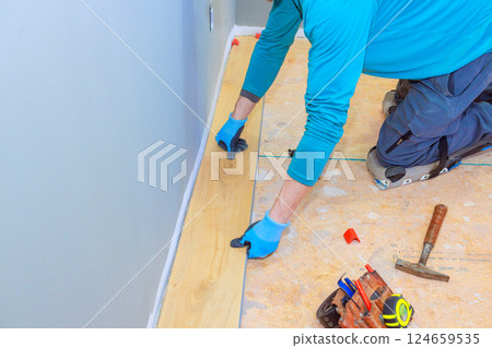 Worker uses tools to install wooden flooring in room space while wearing gloves, kneeling on plywood 124659535