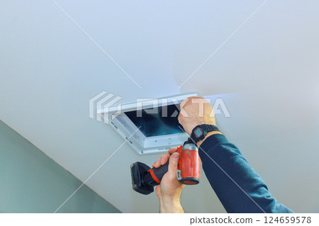 Handyman works on installing ventilation duct cover in ceiling of well lit room, at home repair task. 124659578