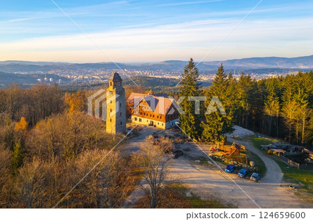 A clear sunny autumn morning highlights the Bramberk lookout tower and adjacent hut nestled in the Jizera Mountains, surrounded by vibrant fall foliage and serene landscapes. 124659600