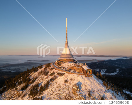 Snow blankets Jested Mountain as the sun rises, casting a warm glow over the landscape. The tower stands tall, surrounded by serene winter beauty and a misty horizon. Snow blankets Jested Mountain as the sun rises, casting a warm glow over the landscape. The tower stands tall, surrounded by serene winter beauty and a misty horizon. 124659608