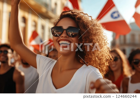 A joyous young woman with curly hair celebrates, wearing sunglasses reflecting a city scene, at a vibrant outdoor event with flags 124660244