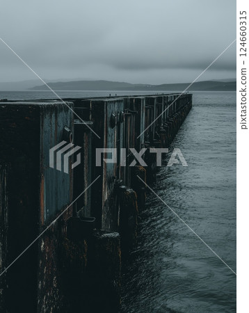 A gloomy seascape featuring a weathered industrial pier stretching into calm waters, with misty mountains in the background A gloomy seascape featuring a weathered industrial pier stretching into calm waters, with misty mountains in the background 124660315