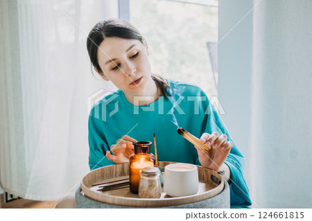 Young woman burning palo santo near a tray with candles and tea in a calm home setting. Sensory mindfulness for daily balance, palo santo ritual, inner calm, grounding therapy 124661815