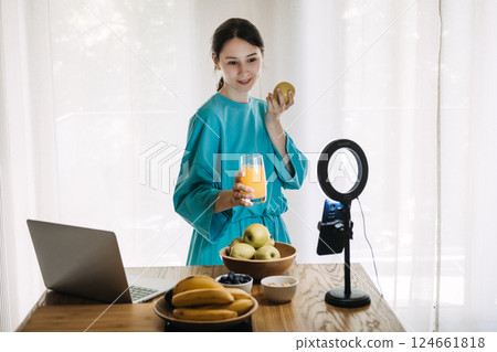 Young white woman in a turquoise robe holds an apple and a glass of juice while filming at home with a ring light and smartphone. Healthy lifestyle, content creator economy, self-branding 124661818