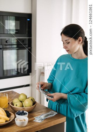 Young woman counting US dollars beside a kitchen counter with fruit and juice. Budgeting in the wellness era, conscious spending, financial self-care, food cost planning 124661861