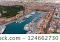 Drone view of Nice, France, showcasing the vibrant harbor filled with yachts and boats, surrounded by colorful historic buildings and the stunning coastline of the French Riviera under a blue sky.  124662730