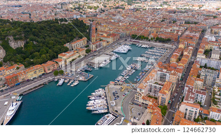 Drone view of Nice, France, showcasing the vibrant harbor filled with yachts and boats, surrounded by colorful historic buildings and the stunning coastline of the French Riviera under a blue sky.  124662730