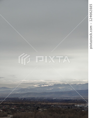 Overcast sky blankets a distant mountain range with a town nestled in the valley. A moody and expansive scene portraying the quiet of nature and settlement below. Overcast sky blankets a distant mountain range with a town nestled in the valley. A moody and expansive scene portraying the quiet of nature and settlement below. 124662885