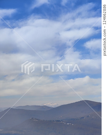 Snowy mountaintops rise above layers of dark hills and scattered villages. The contrast between fog, sky, and mountains creates a dramatic natural composition. 124662886