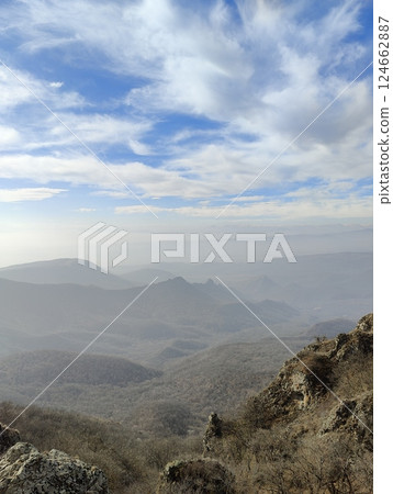View from a high point overlooking a series of forested hills and distant mountain peaks. The partly cloudy sky adds texture to the layered mountain landscape. 124662887