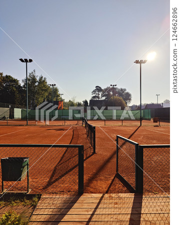 Early morning sunlight casts long shadows over a clay tennis court surrounded by fencing and trees. The peaceful setting and warm light create a tranquil atmosphere. Early morning sunlight casts long shadows over a clay tennis court surrounded by fencing and trees. The peaceful setting and warm light create a tranquil atmosphere. 124662896