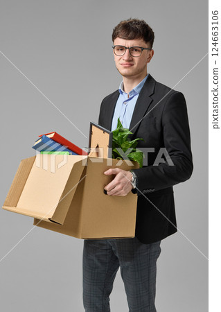 Young businessman with serious expression holding cardboard box with books and plant. Concept of unexpected job change Young businessman with serious expression holding cardboard box with books and plant. Concept of unexpected job change 124663106