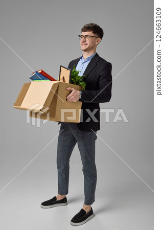 Happy young professional holding box full of office items against grey background, showing new beginning, career change. Happy young professional holding box full of office items against grey background, showing new beginning, career change. 124663109