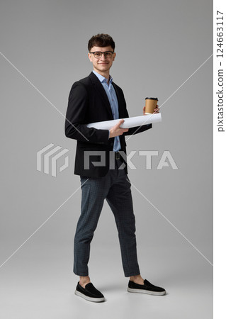 Full length portrait of confident young businessman in suit holding coffee and blueprint against grey background. Concept of ambition Full length portrait of confident young businessman in suit holding coffee and blueprint against grey background. Concept of ambition 124663117