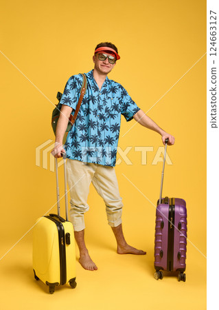 Full body photo of happy young man barefoot in tropical shirt holding two suitcases, smiling confidently against yellow background. Full body photo of happy young man barefoot in tropical shirt holding two suitcases, smiling confidently against yellow background. 124663127