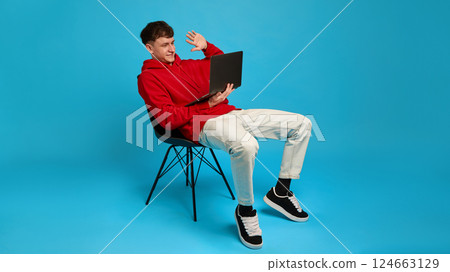Relaxed young man on chair waving to laptop screen during online conversation, against blue background. Concept of distance work Relaxed young man on chair waving to laptop screen during online conversation, against blue background. Concept of distance work 124663129