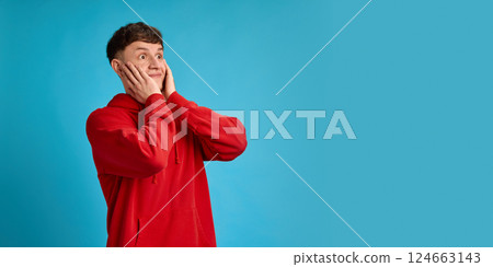 Funny surprised man in red hoodie holding face with both hands looking up against blue studio background. Concept of shock 124663143