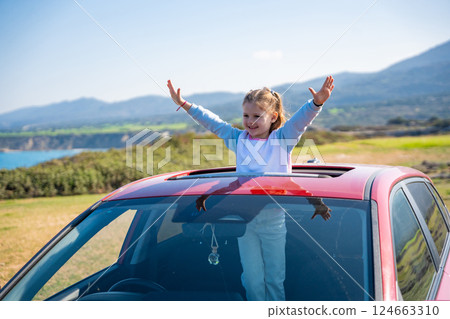 Little girl joyfully sticks out of car sunroof with raised hands against the backdrop of mountains in Northern Cyprus. The image symbolizes freedom, adventure, and the excitement of travel with child Little girl joyfully sticks out of car sunroof with raised hands against the backdrop of mountains in Northern Cyprus. The image symbolizes freedom, adventure, and the excitement of travel with child 124663310