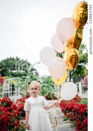 Young child in white dress holding colorful balloons in a vibrant floral garden setting 124663762