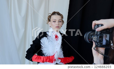 Professional photographer capturing fashion model wearing red gloves, posing with white feather against monochromatic studio backdrop Professional photographer capturing fashion model wearing red gloves, posing with white feather against monochromatic studio backdrop 124666203