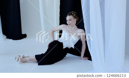 Fashion model wearing a white top and black pants is posing on the floor in a studio with black and white curtains, creating a contrasting and stylish atmosphere Fashion model wearing a white top and black pants is posing on the floor in a studio with black and white curtains, creating a contrasting and stylish atmosphere 124666213