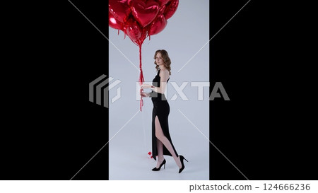 Sophisticated woman wearing black dress, gripping red heart shaped balloons, celebrating valentine's day elegantly against neutral studio background 124666236