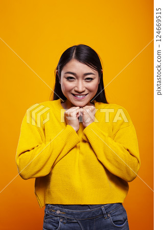 Cheerful woman looking at camera with her hands placed gently near her chin, exuding warmth and friendliness. Smiling asian lady posing against an isolated backdrop and resting her jaw on her fists. 124669515