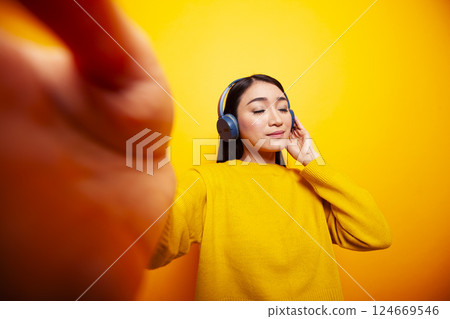 Asian vlogger standing against isolated backdrop and listening to music. Young woman with headphones, recording daily vlog and holding a camera, with relaxed expression in front of yellow background. Asian vlogger standing against isolated backdrop and listening to music. Young woman with headphones, recording daily vlog and holding a camera, with relaxed expression in front of yellow background. 124669546