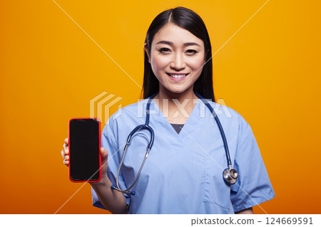Young doctor in medical uniform, holding smartphone with black screen against a yellow background, gazing at camera. Portrait of asian woman in nurse scrubs, grasping mobile device with blank display. 124669591