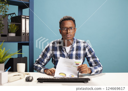 Businessman seated at desk with paperwork, suspicious of the startup project results, having confused expression. POV of male finance analyst checking company accounts on computer, unsure of expenses. 124669617