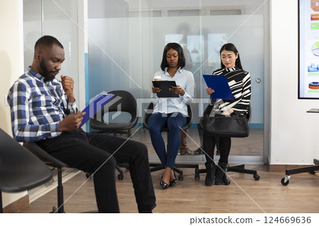 Small group of applicants in office lobby, reading company internal regulations and preparing for employment meeting. Multiethnic unemployed people sitting in queue for job interview process. 124669636