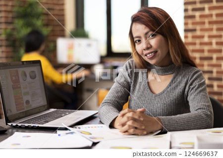 Asian businesswoman sits confidently at her office desk, smiling at the camera, ready to tackle projects. Portrait of female professional prepared for busy workday, surrounded by paperwork and laptop. 124669637