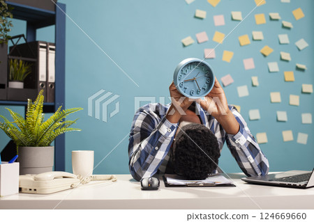 Exhausted male manager resting his head on table and carrying an analogue clock, counting down to end of workday. African american businessman collapsed on office desk, holding alarm clock above him. 124669660