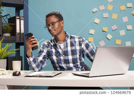 Young male freelancer sitting at his desk and looking at his mobile device. African american consultant holding his smartphone, reading text messages as he takes a break from his tasks. 124669679