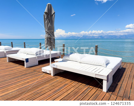 Sunbeds on wooden pier on shore of sea in summer landscape. Sunchairs on wooden deck by the ocean. 124671335