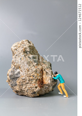 A tiny figurine of a man pushing a massive rock, symbolizing perseverance, struggle, and determination. 124671512