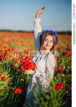 Young woman wearing blue flower crown and white dress holding poppy bouquet with closed eyes and raised arm in a poppy field Young woman wearing blue flower crown and white dress holding poppy bouquet with closed eyes and raised arm in a poppy field 124674167