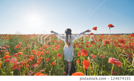 Young woman with long blond hair and a flower crown walks through a vibrant poppy field at sunset, embracing the beauty of nature 124674191