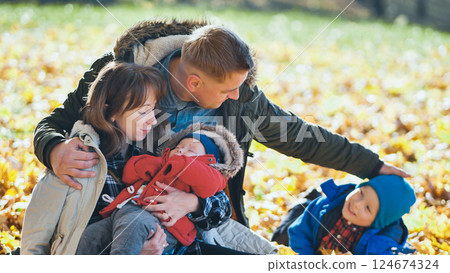 Family cuddling newborn, caressing toddler among fallen leaves during golden autumn sunlight in peaceful park setting Family cuddling newborn, caressing toddler among fallen leaves during golden autumn sunlight in peaceful park setting 124674324