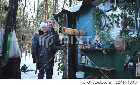 Homeless couple standing beside ramshackle shelter, weathering winter landscape with quiet determination amid challenging circumstances 124674339